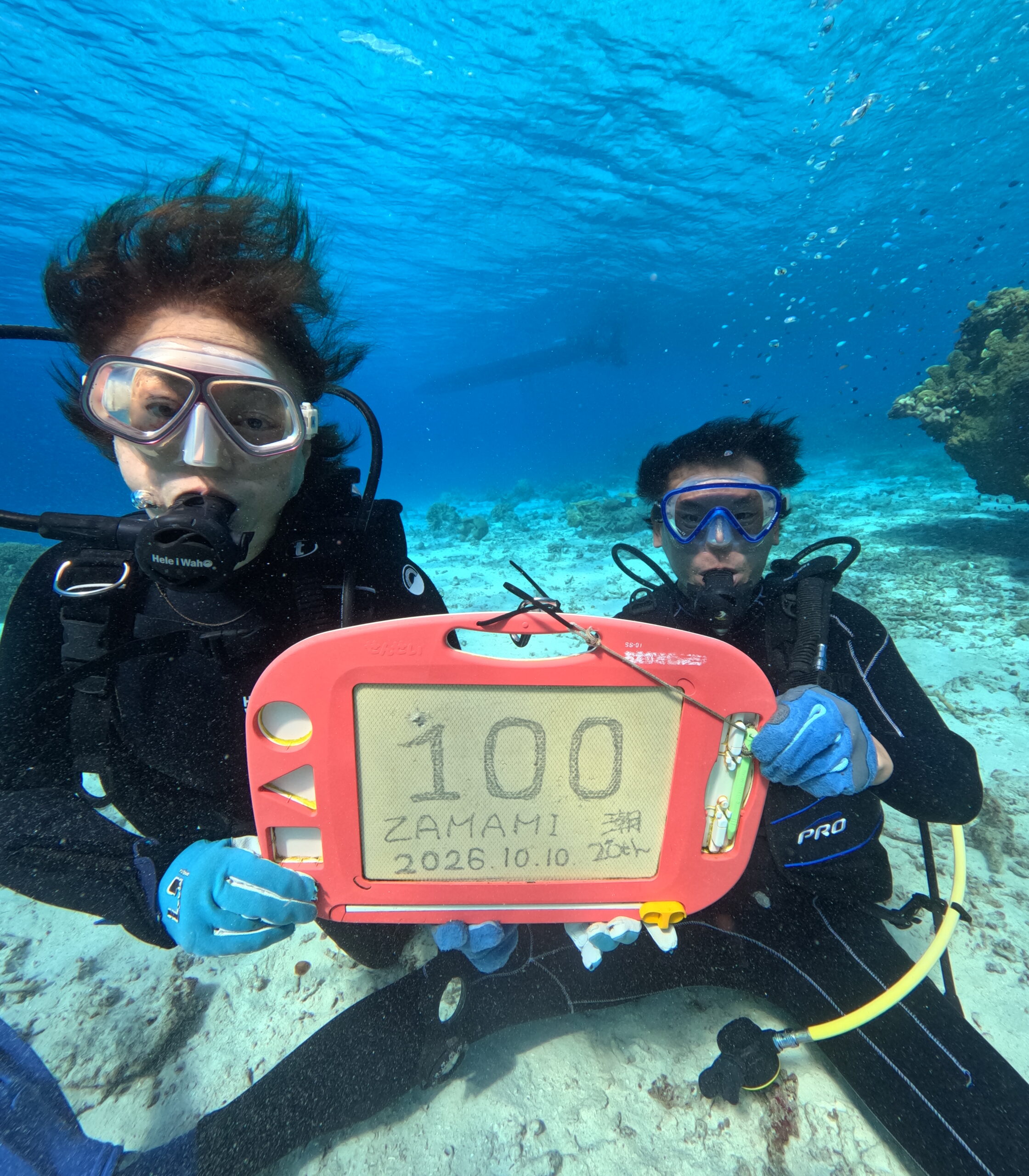 座間味のケラマブルーの海で100本記念のスレートを持つご夫婦ダイバー。透明度30mの美しい海で迎えた記念ダイビング。